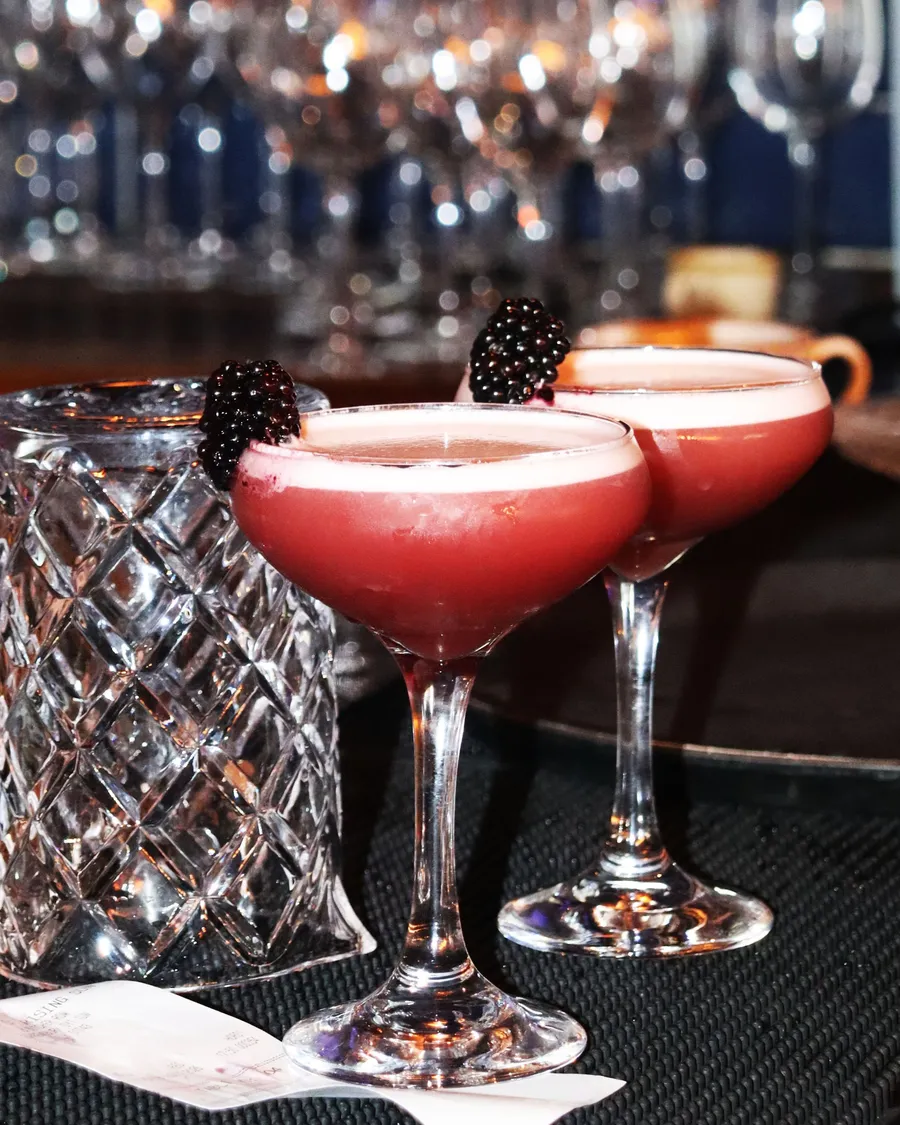 Two pink sours garnished with blackberries on the bar